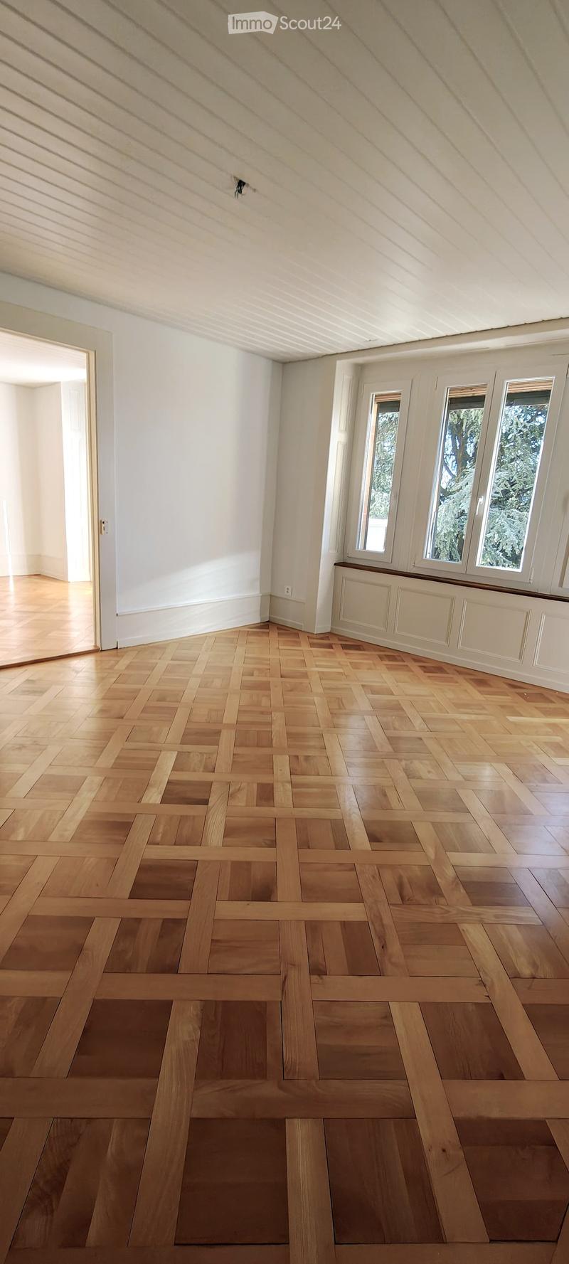 Empty room with wooden herringbone flooring, white walls, and large windows with white frames. The ceiling has exposed wooden beams and a skylight. There is a radiator under the windows and an open doorway leading to another space.