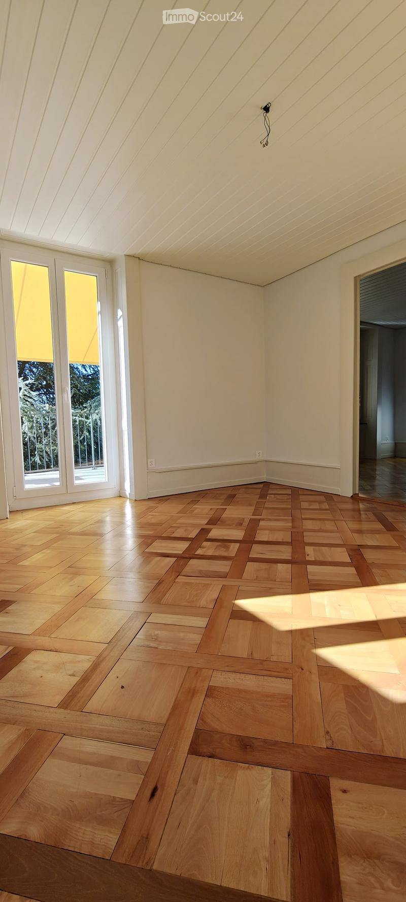 Empty room with wooden herringbone flooring, white walls, and large windows letting in natural light.