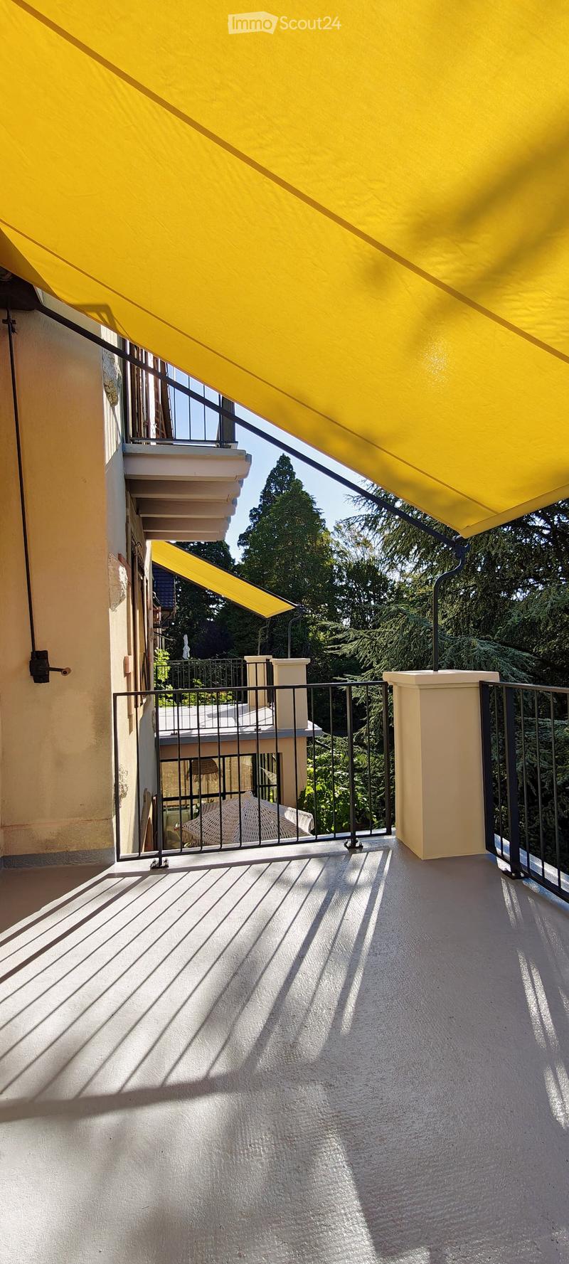Balcony with a yellow awning, black metal railing, and a view of greenery.