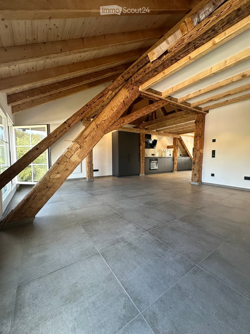 Open-plan space with exposed wooden beams and pillars, grey tiled floor, modern kitchen area with cabinets, refrigerator, and oven, large windows with a view of greenery.