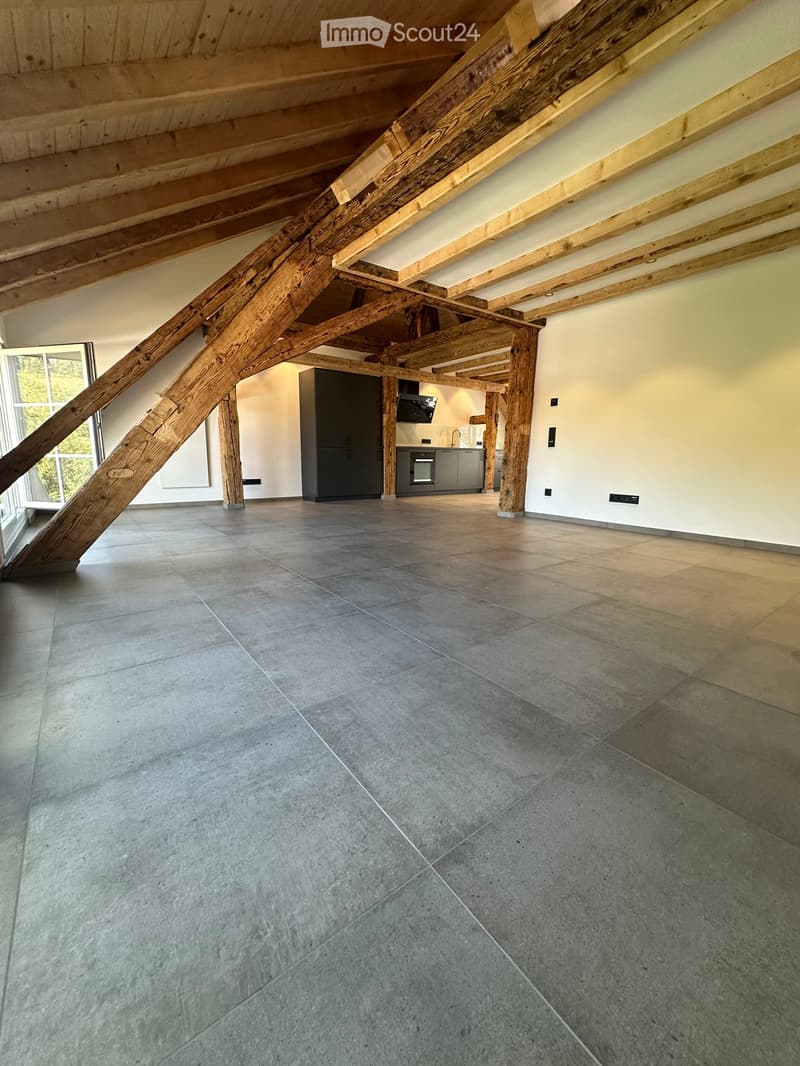 Open, high-ceilinged room with exposed wooden beams and grey tiled flooring. Kitchen area visible in the background with cabinets and appliances.