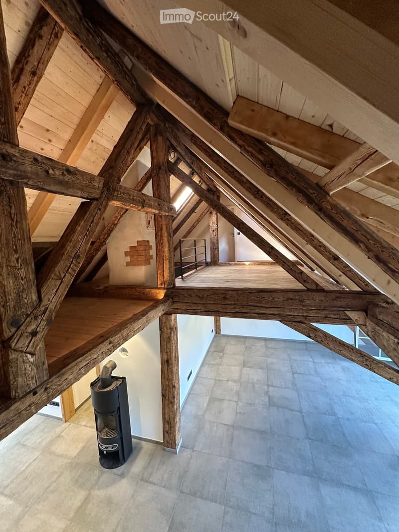 Attic hallway with exposed wooden beams, concrete floor, and a black wood-burning stove on the left side.