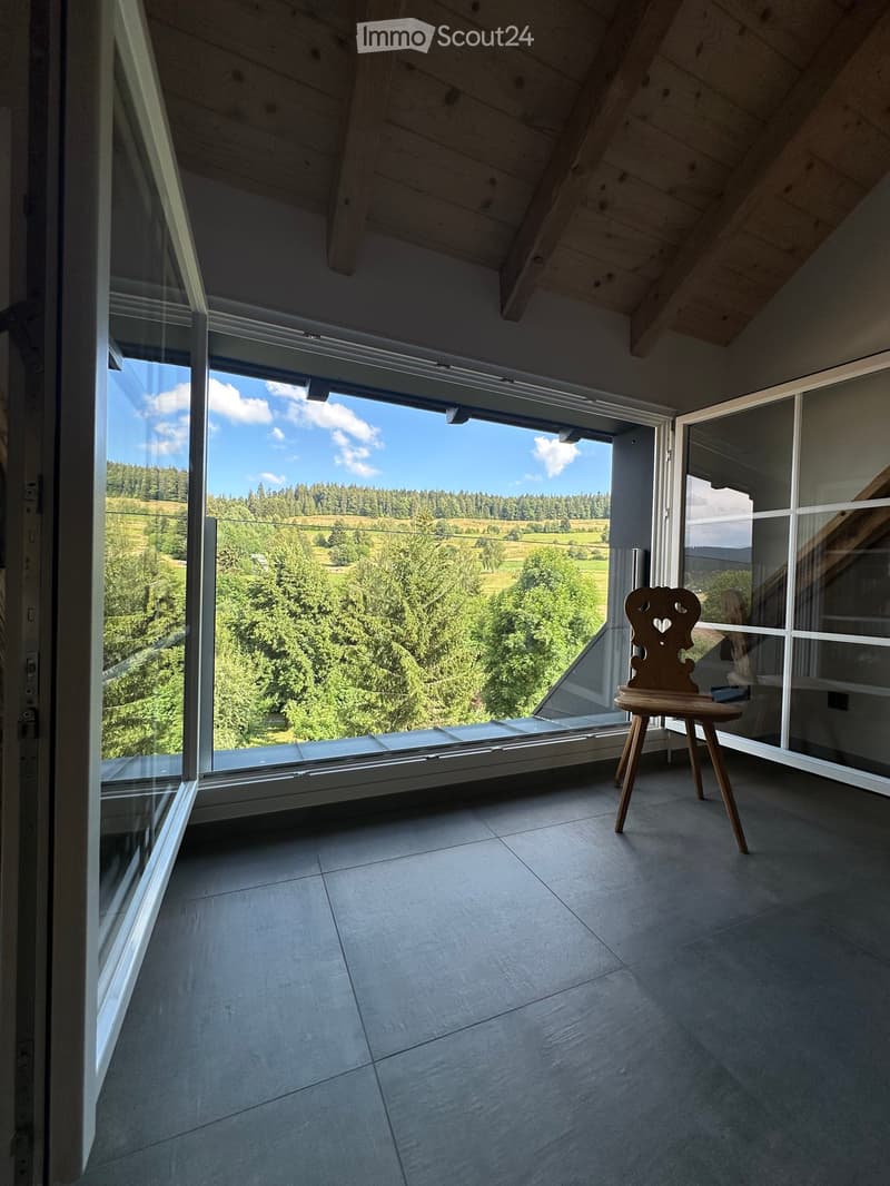 Modern room with large windows offering a scenic view of a green landscape, featuring a single wooden stool.