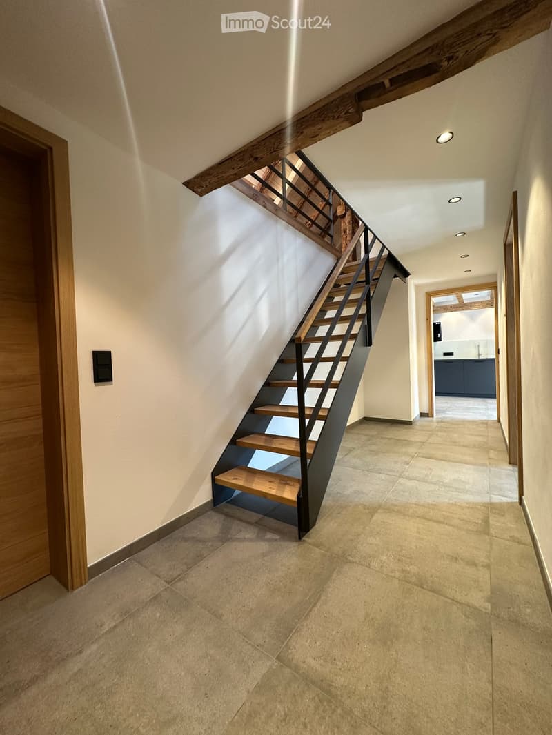 Modern hallway with a wooden staircase featuring a black metal railing, beige tiled floor, and recessed ceiling lights. A doorway leads to a kitchen area with dark cabinets and a white countertop.
