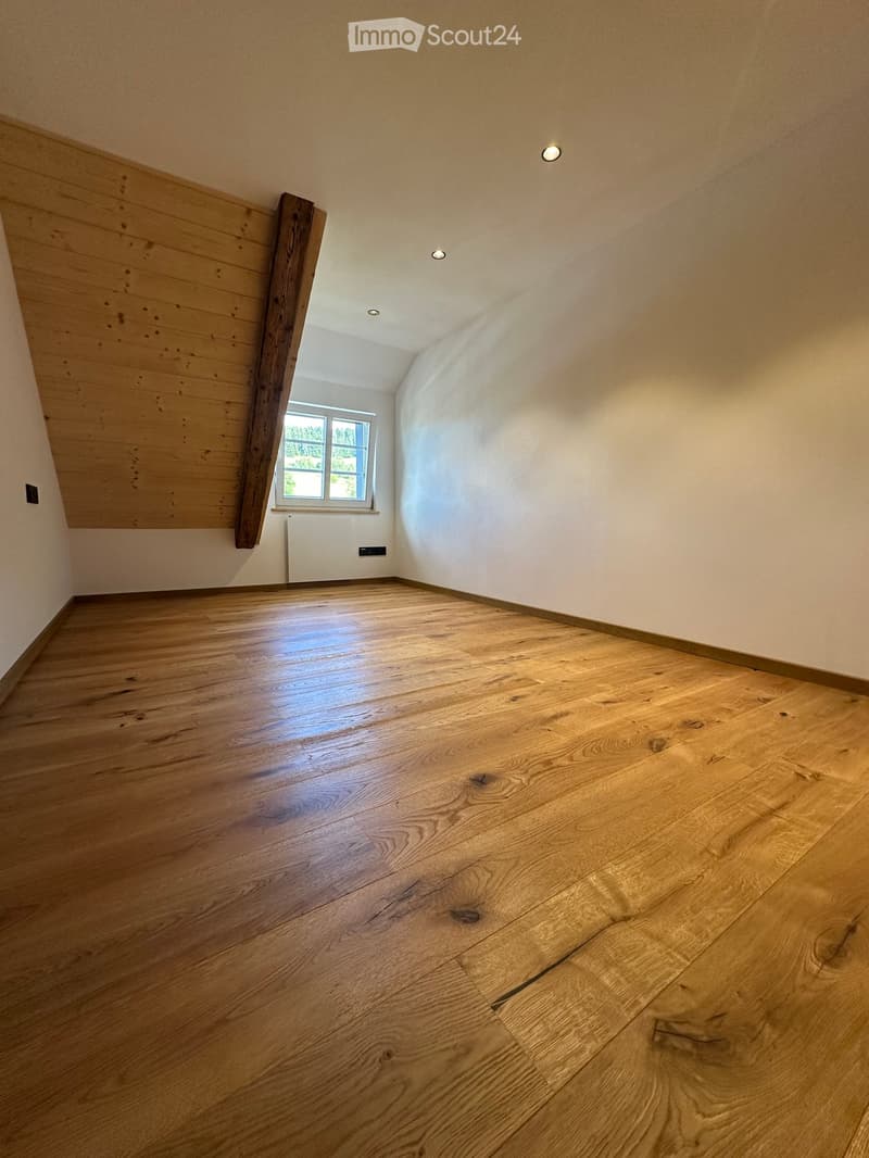 Empty room with wooden flooring, white walls, and a single window letting in natural light. The room features recessed ceiling lights and a radiator under the window.