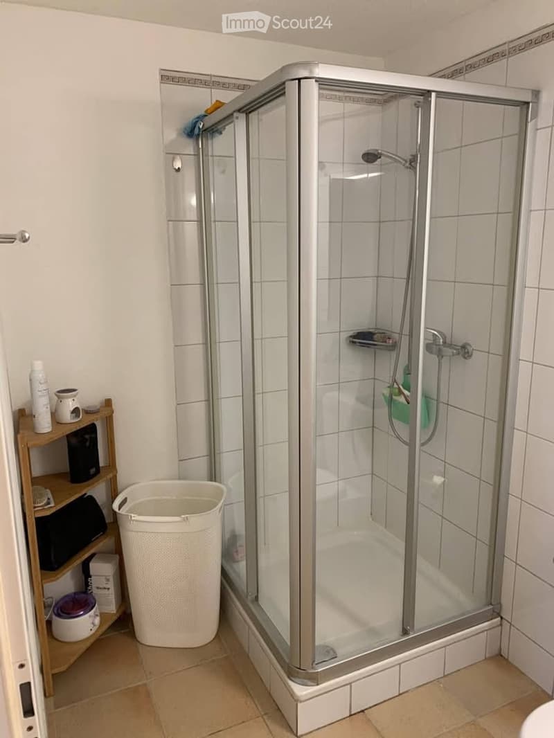 Modern bathroom with a glass-enclosed shower stall, tiled walls, a wooden shelf holding various items, and a white trash bin on the floor.