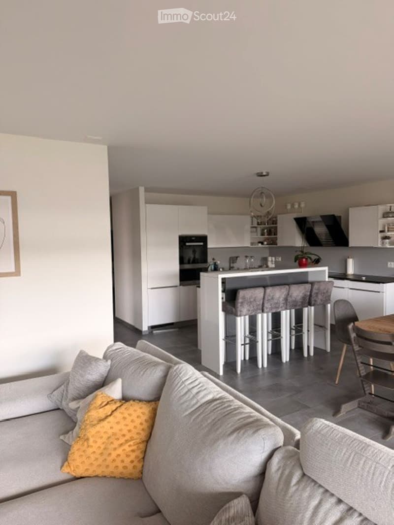 Modern open-plan living and dining area with a grey sofa, yellow throw pillow, kitchen island with four stool chairs, white cabinets, and an oven. A framed picture hangs on the wall.