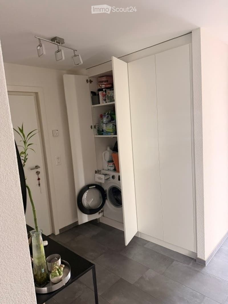 A modern, compact laundry room with white cabinets, a built-in washing machine, and a small table with decorative items.
