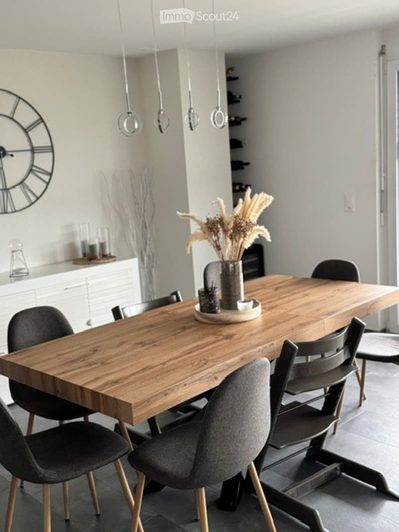 Modern kitchen with a long wooden dining table surrounded by grey chairs, a decorative clock on the wall, and a vase with dried flowers on the table.