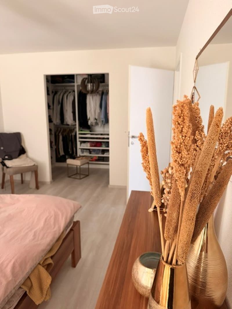 Modern bedroom with light wooden flooring, a neatly made bed, a small chair, a large mirror, and decorative vases on a wooden console table. An open closet with organized clothing and accessories is visible in the background.