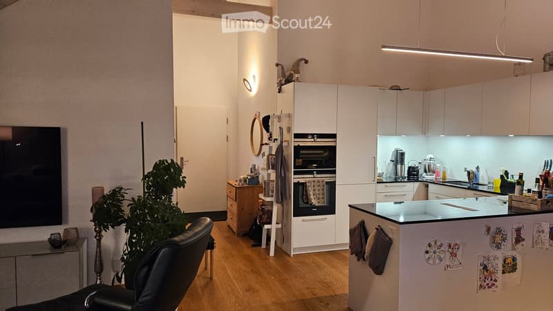 Modern, open-plan living and kitchen area with light wood flooring, white cabinetry, and a central island. Features a black leather sofa, a small wooden table with chairs, a built-in oven, and a refrigerator. The kitchen area includes a sink, faucet, and