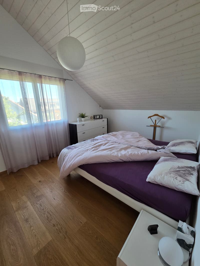 Cozy attic bedroom with wooden floors, slanted wooden ceiling, large window with sheer curtains, bed with pink bedding, nightstand, lamp, and small dresser.