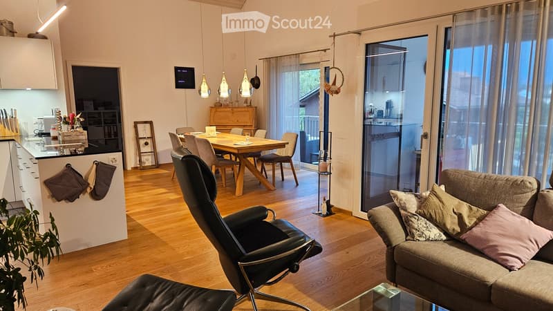 Open-plan living and dining area with wooden flooring, a grey sofa with pillows, a black leather chair, a wooden dining table with chairs, a modern kitchen counter, pendant lights, and a screen on the wall.