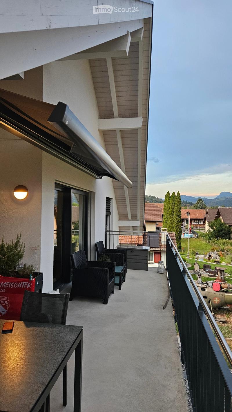 Balcony with a table, chairs, and a small stool, overlooking a scenic landscape with houses and mountains.