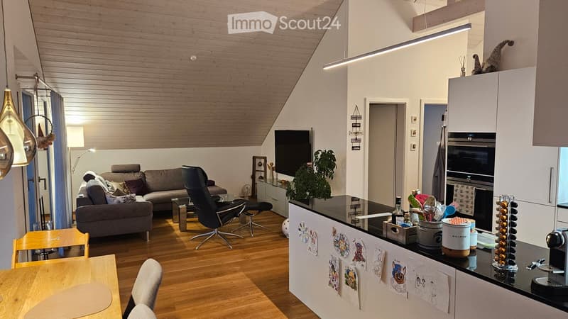 A modern, open-plan living and kitchen area with wooden flooring, white walls, and a slanted wooden ceiling. The living area features a grey sofa, a black leather chair, a small glass coffee table, and a wooden dining table with chairs. The kitchen area i