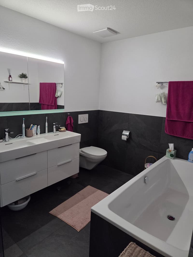 Modern bathroom with a white sink, toilet, bathtub, and large mirror. Features dark tiles, a pink bath mat, and a towel hanging on the wall.