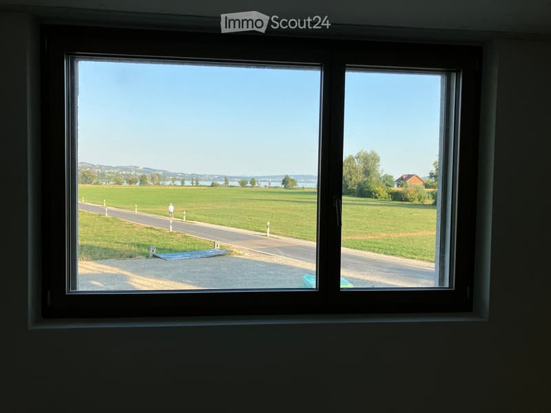 View from a window showing a grassy field, road, and distant lake with hills.