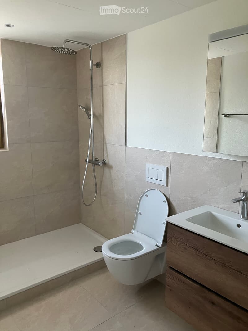 Modern bathroom with beige tiled walls and floor, white toilet, sink with wooden cabinet, and a shower area with a handheld showerhead.