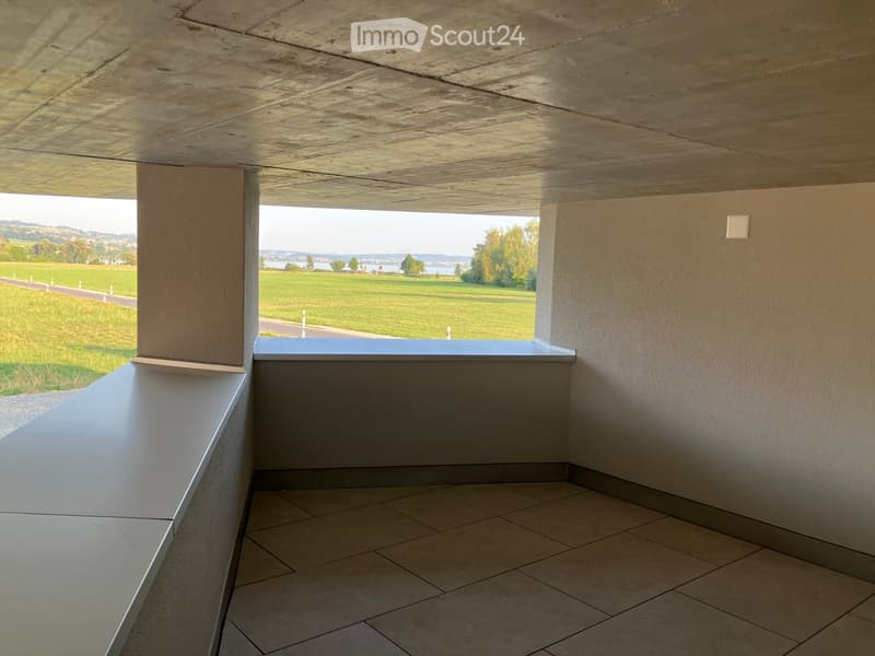 Modern balcony with tiled floor, concrete seating area, and expansive view of a green field and water body.