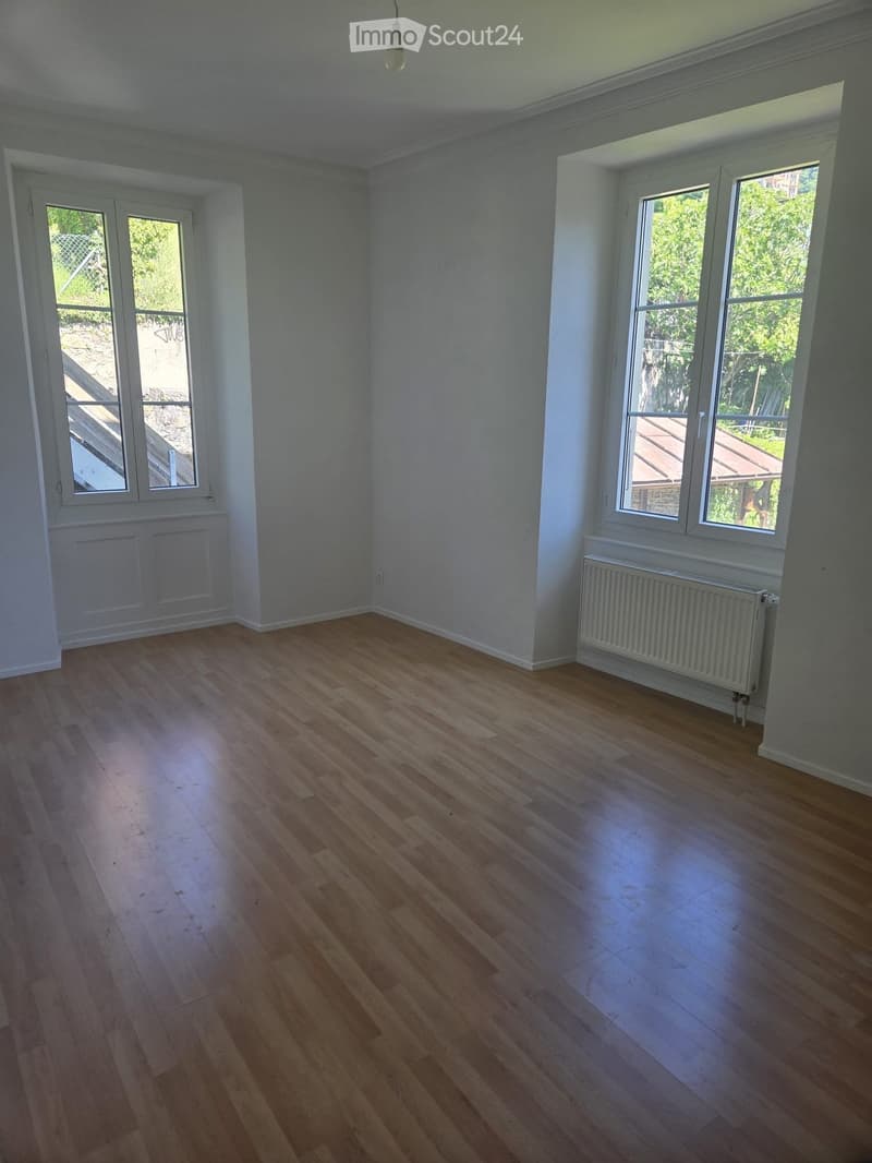 Empty room with wooden flooring, white walls, large windows, radiator, and a hanging light fixture.