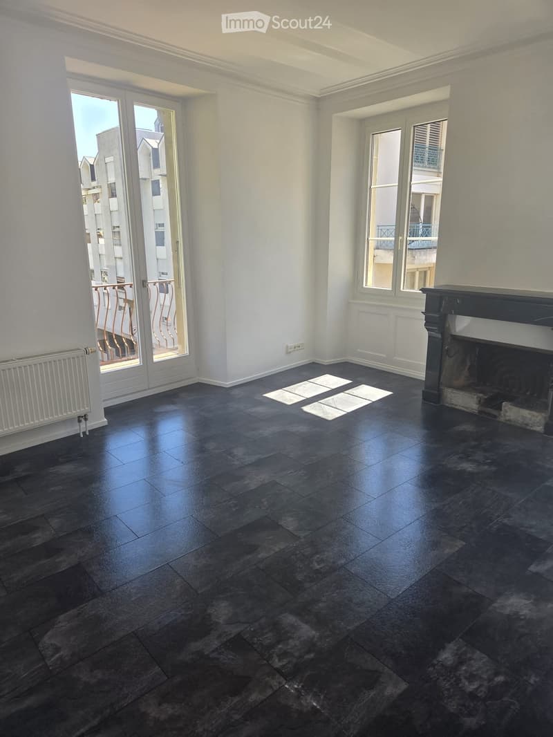 Empty room with dark tiled flooring, white walls, large windows, and a fireplace. Features include a radiator, a balcony door, and a view of the building exterior.
