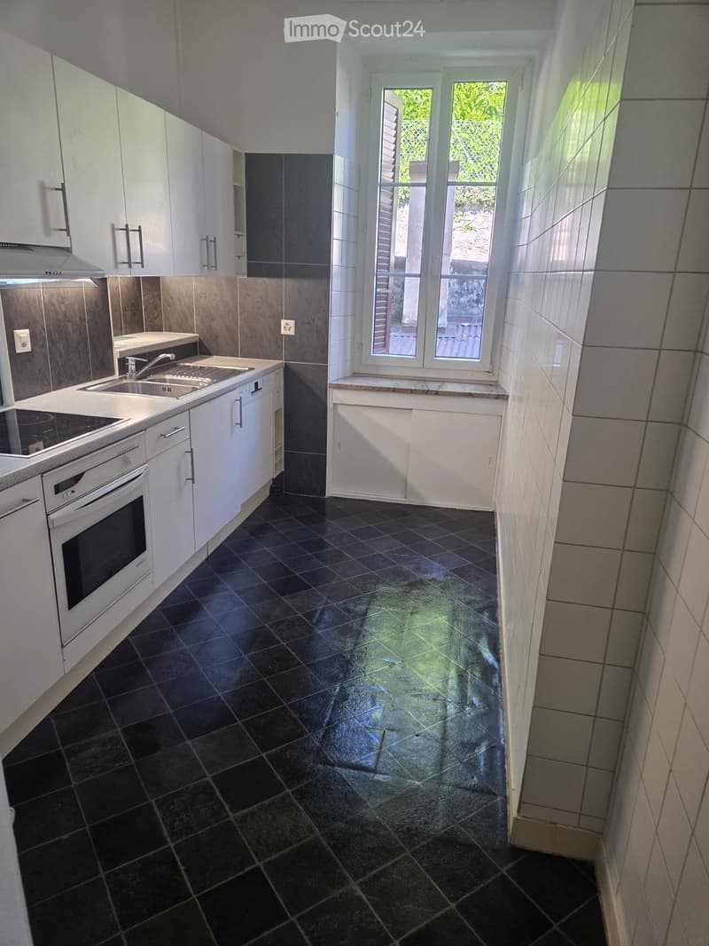 Modern kitchen with white cabinets, black tiled floor, stainless steel sink, oven, and dishwasher. Large window with view of greenery.