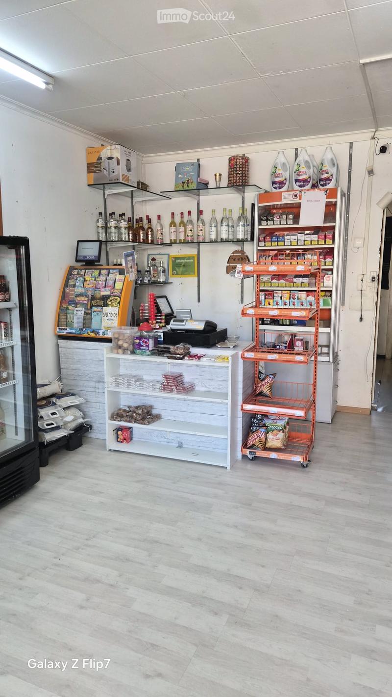 A well-lit convenience store interior with a clean, modern design. The space features a central counter with a cash register, shelves stocked with various products, and a refrigerator on the left side. The floor is made of light-colored tiles, and the wal