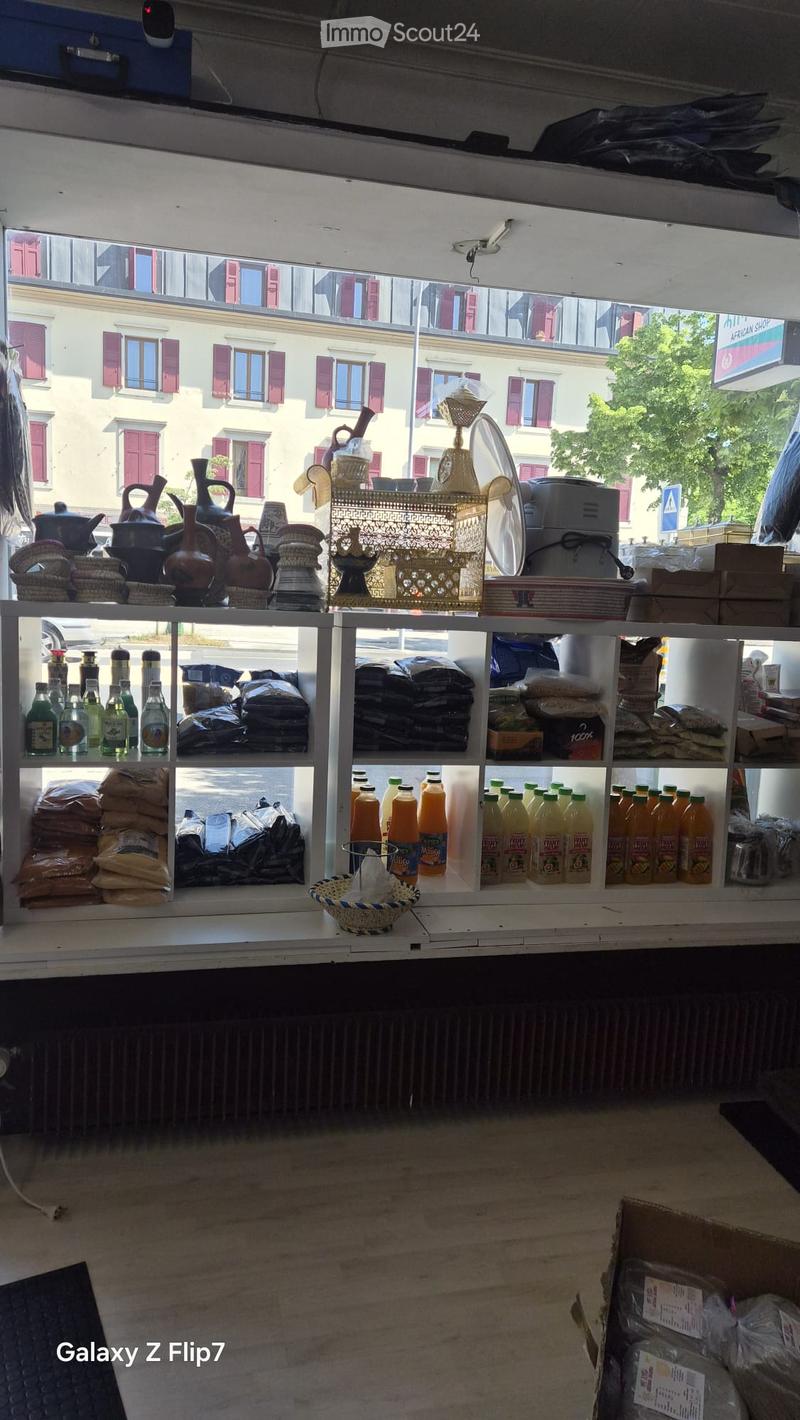 Shop interior with shelves displaying various items including bottles, teapots, vases, decorative items, and orange juice bottles. Visible through a large window showing a building with red shutters in the background.
