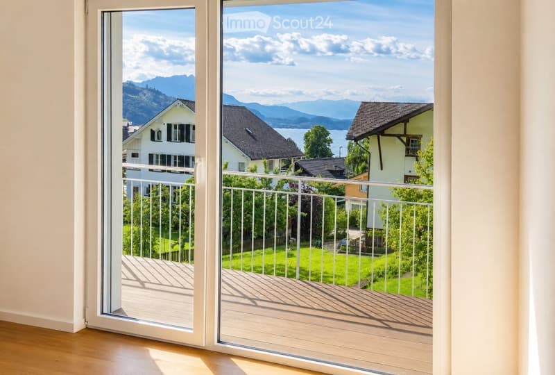 Open sliding glass doors lead to a wooden deck with white railing, overlooking a scenic view of mountains, a lake, and neighboring houses.
