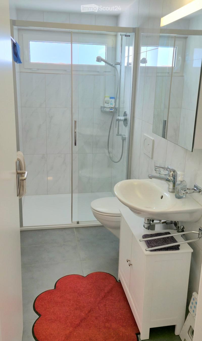 white shower with shower head, white toilet, white sink, white shelves, red floor mat, wall-mounted mirror