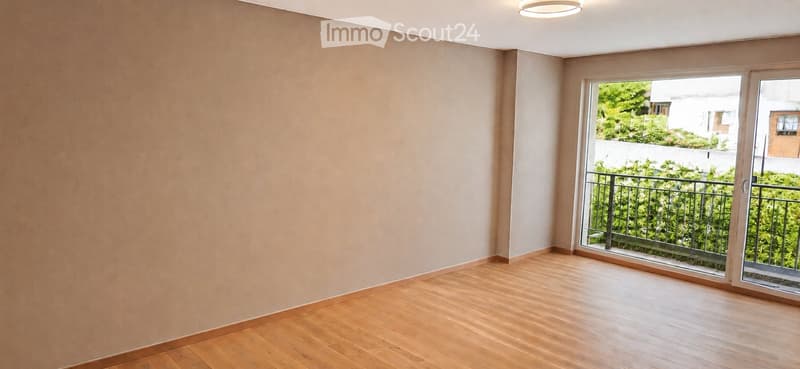 Empty room with wooden flooring, light-colored walls, and a large sliding glass door leading to a balcony with a metal railing and greenery visible outside.