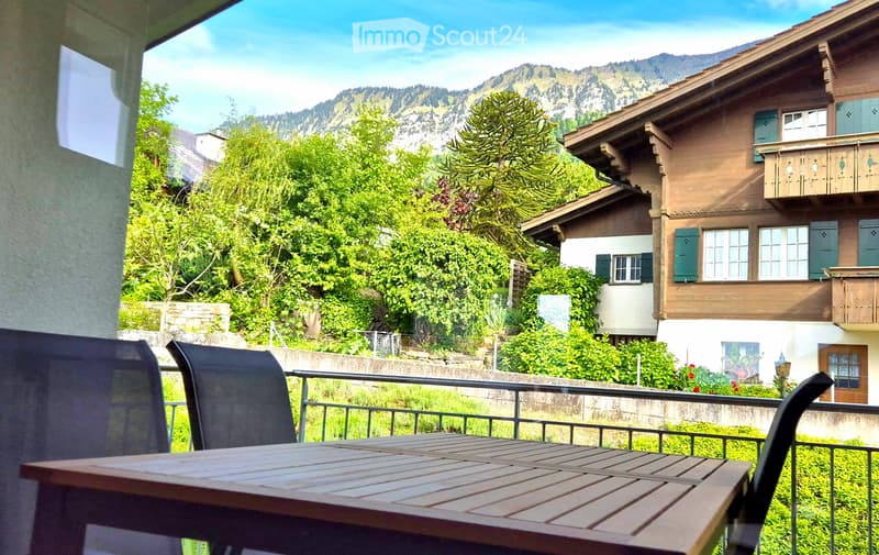 Balcony with table and chairs, surrounded by garden, distant view of mountains.