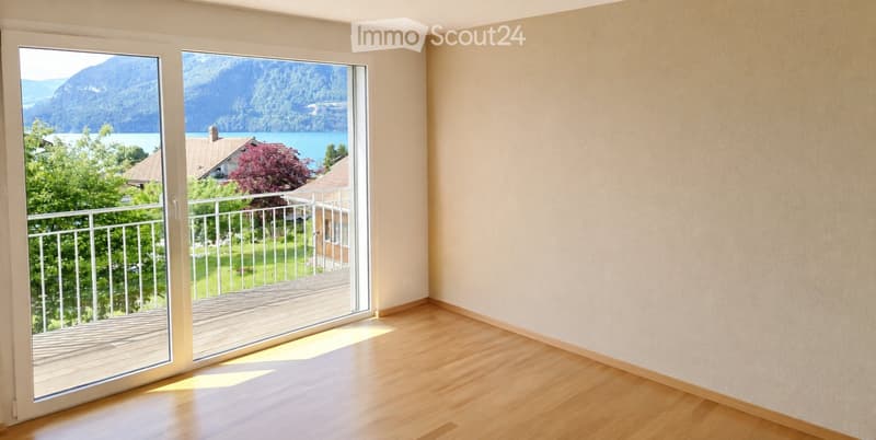 Empty room with wooden flooring, large windows offering a scenic view of a lake and mountains, and a balcony accessible through the windows.