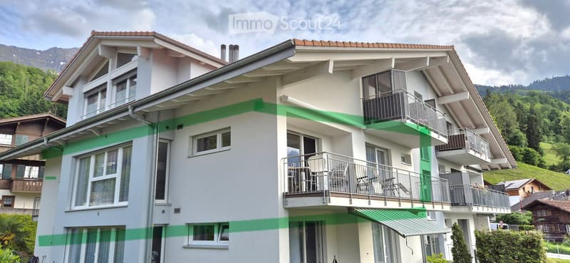 Alpine-style white house with brown roof, multiple balconies, green stripe painting, surrounded by lush greenery