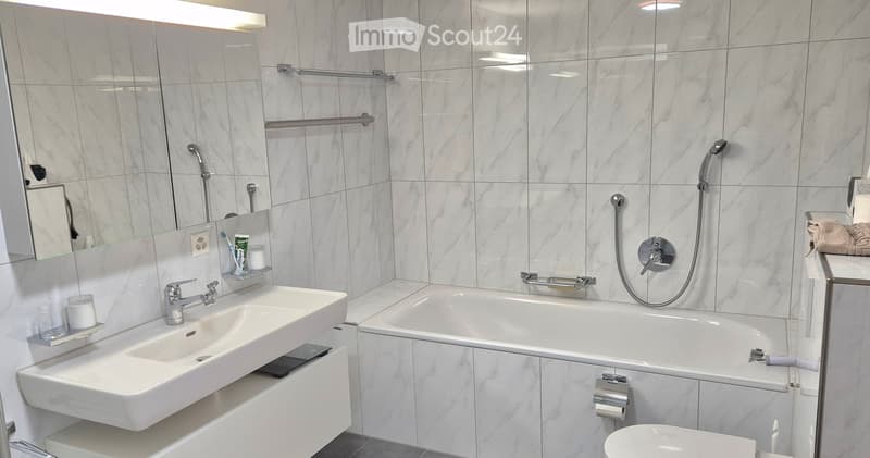 modern bathroom with white marble tiles, bathtub, shower head, sink, mirror, wall-mounted faucet, glass cup, tissue holder, soap dispenser, toilet