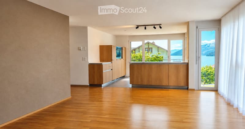 Modern kitchen with light wooden cabinets, stainless steel oven, and large windows offering a scenic view of a lake and mountains.