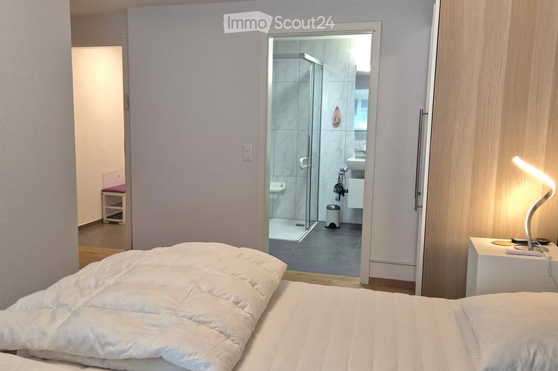 white bedding, open doorway to bathroom, modern lighting, small table with lamp, large mirror on wall
