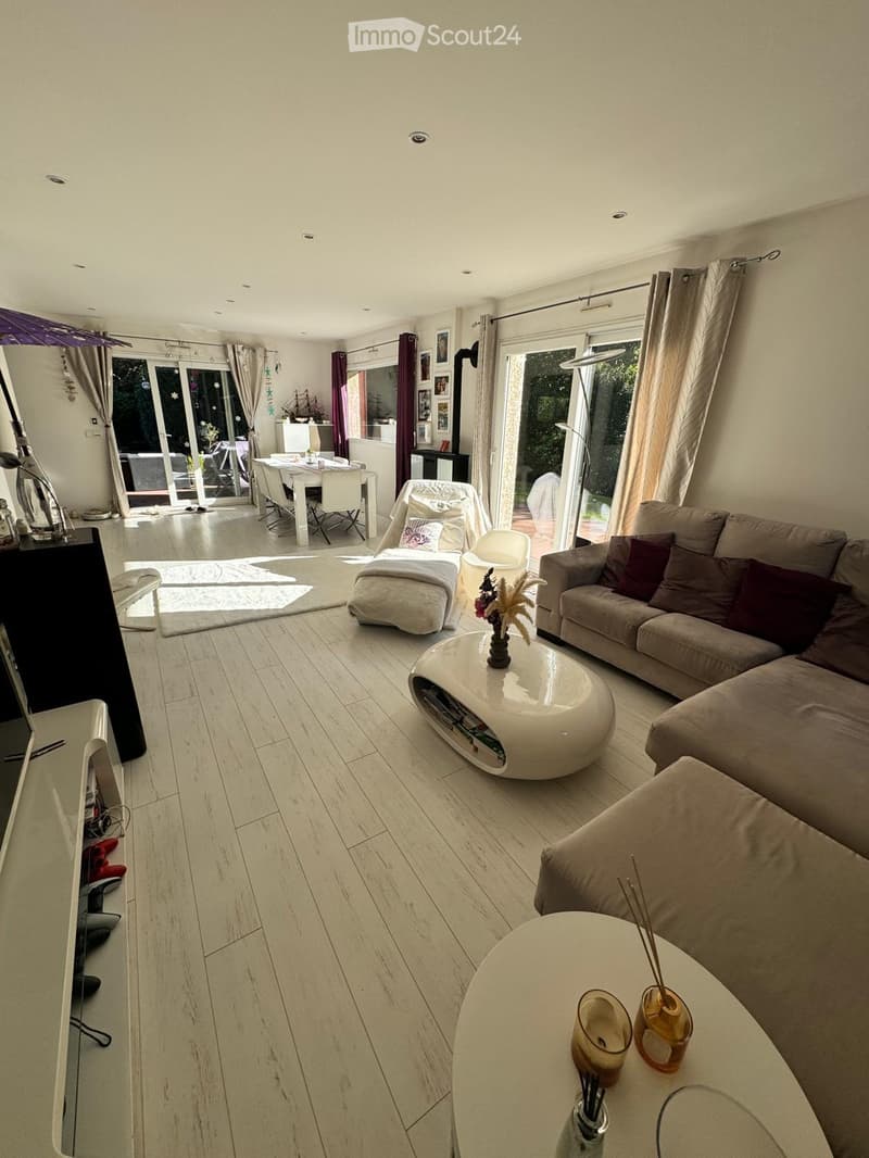 Modern living room with light wood flooring, white walls, and large windows. Features a beige sectional sofa, white coffee table, ottoman, dining area with white table and chairs, and decorative vases.