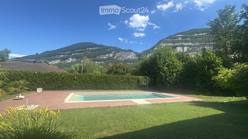 Outdoor garden area with a swimming pool, wooden deck, lush greenery, and a mountain view in the background.