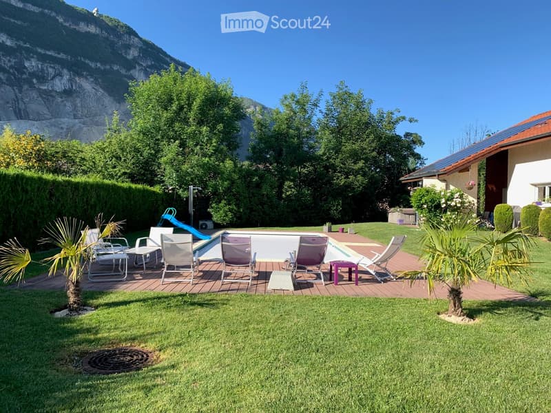 Well-maintained garden with a swimming pool, lounge chairs, and a slide, surrounded by greenery and a mountain backdrop.