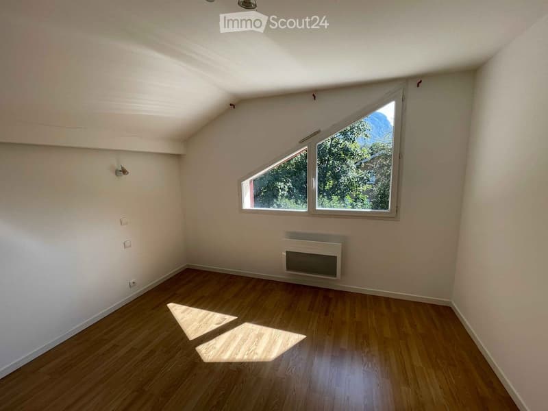 Empty room with wooden flooring, white walls, and large windows offering a view of greenery. The room features a radiator and a simple ceiling design.