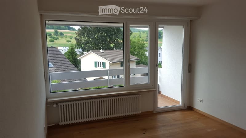 Empty room with wooden flooring, large windows, radiator, and sliding door leading to a balcony with a view of a green landscape and houses.