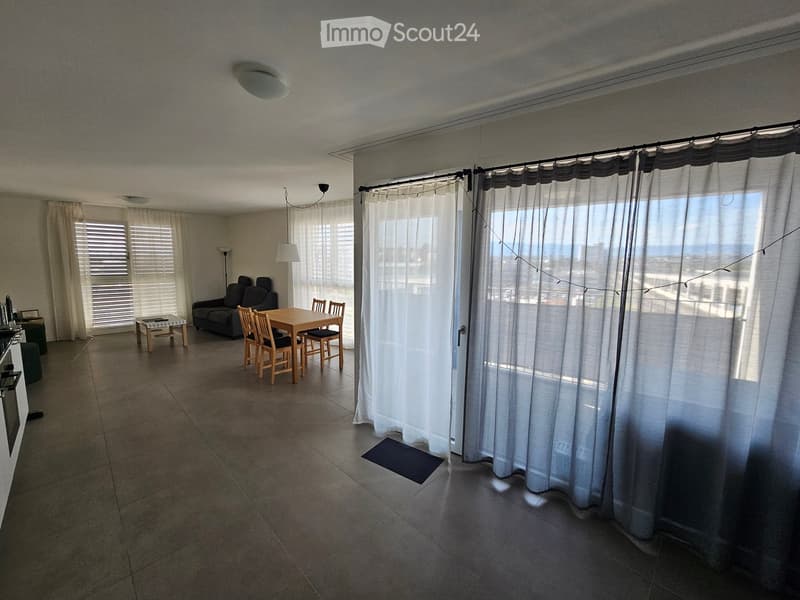 Bright living space with tiled floor, large windows with curtains, sofa, dining table with chairs, and a small stool near the kitchen area.