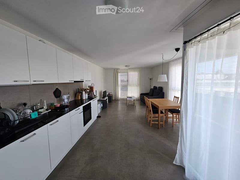 Open-plan living and dining area with a kitchen on the left, featuring white cabinets, a black countertop, and various kitchen items. A wooden dining table with chairs is positioned near a sofa and a small stool, with large windows and white curtains allo