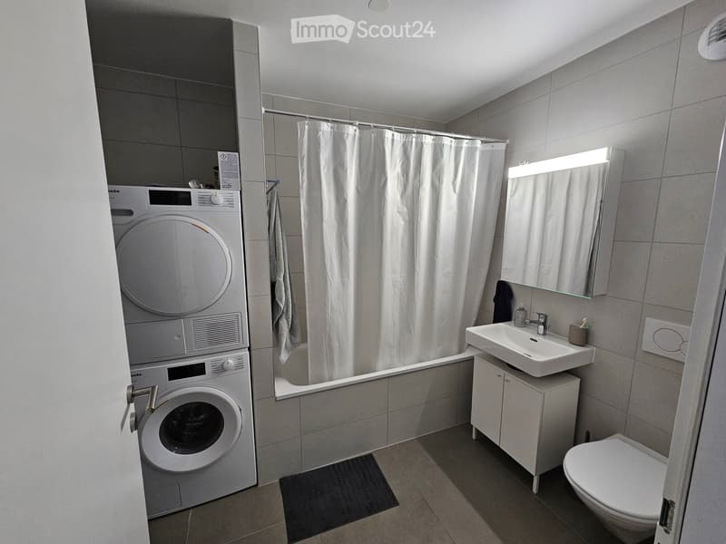 Modern bathroom with a washing machine and dryer unit, bathtub with shower curtain, sink with mirror, and toilet.