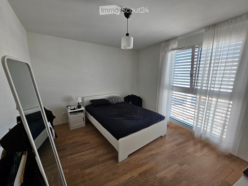 Bedroom with a white bed, dark bedding, nightstand, mirror, and a hanging pendant light. The room has wooden flooring and large windows with blinds.