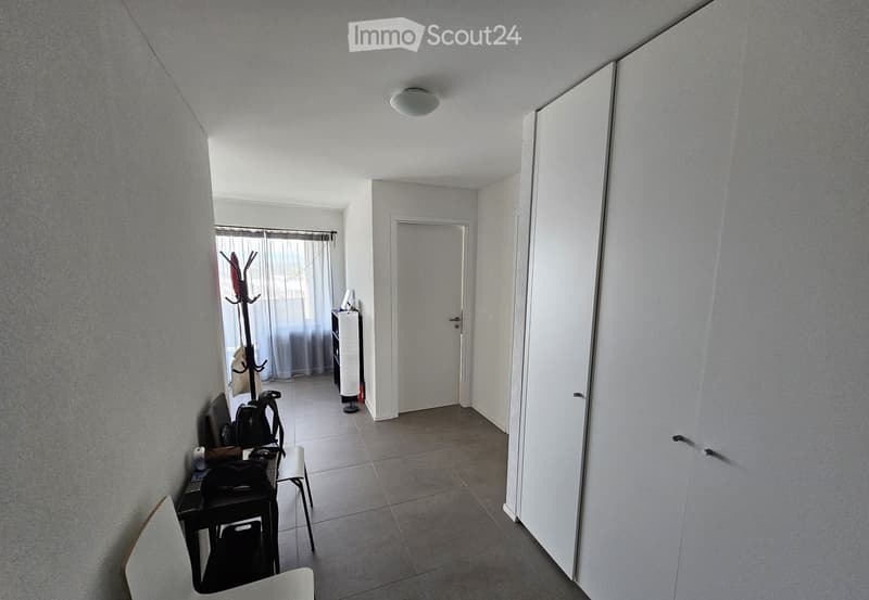 A narrow hallway with tiled flooring, white walls, and a closed door at the end. A small table with a backpack and other items sits near the wall, and a coat rack with a coat is visible in the background.