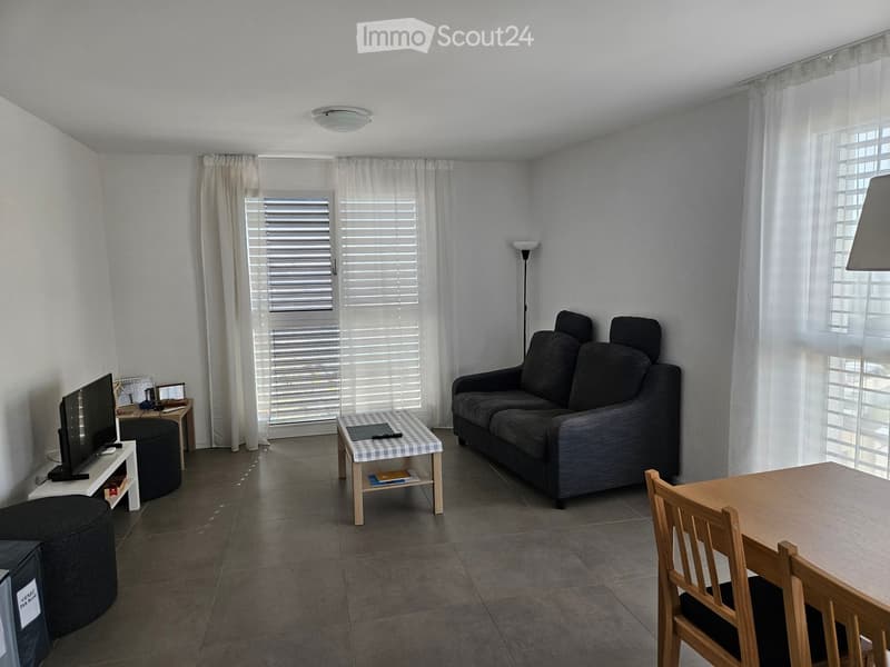 Living room with a grey sofa, coffee table, dining table, chairs, and a TV stand with a screen. The room has large windows with white curtains and a tiled floor.