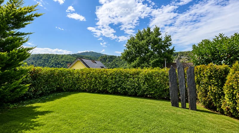 A well-maintained garden with a neatly trimmed hedge, a wooden gate, and a small yellow house in the background, surrounded by lush greenery and trees under a clear blue sky.
