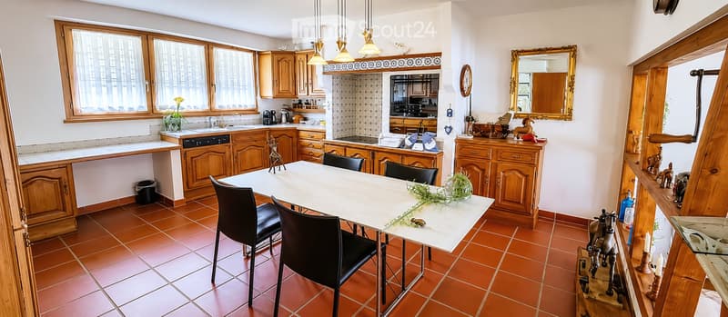 Bright kitchen with wooden cabinets, tiled floor, and a dining area featuring a white table and black chairs. The kitchen includes a sink, dishwasher, and a decorative mirror on the wall.